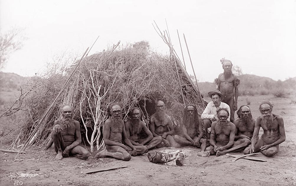 Fig. 11. Une communauté aborigène devant une habitation. Longues sagaies de chasse et de guerre appuyées contre la hutte. Au sol des boomerangs. (http://carreephemere.blogspot.ch/2017/03/architecture-du-monde-laustralie.html)